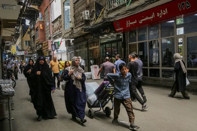 نمایی از یک خیابان شلوغ در تهران با مغازه‌های متعدد. زنان و مردان در خیابان در رفت‌وآمد هستند و در پیش‌زمینه تصویر، پسری جوان در حال کشیدن یک چرخ‌دستی دیده می‌شود

