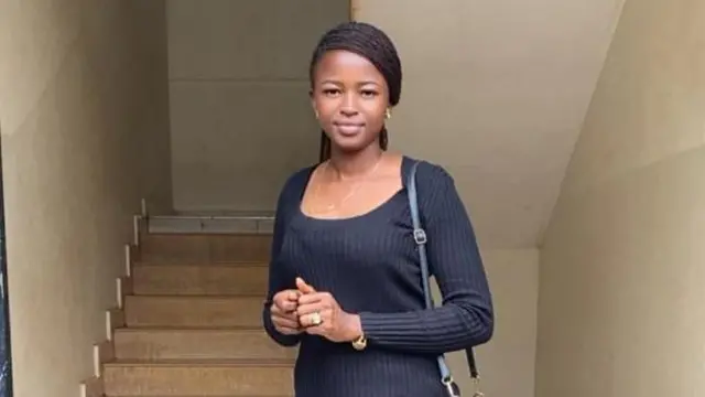 Noella Mukumbi, de pé em frente a uma escada, com blusa e calça preta e carregando uma bolsa da mesma cor, sorri para a câmera