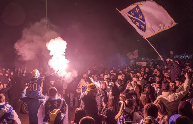 Толпа людей, празднующих победу Боснии над Италией. Многие завернуты в старые флаги Боснии с желтыми геральдическими лилиями на синем фоне