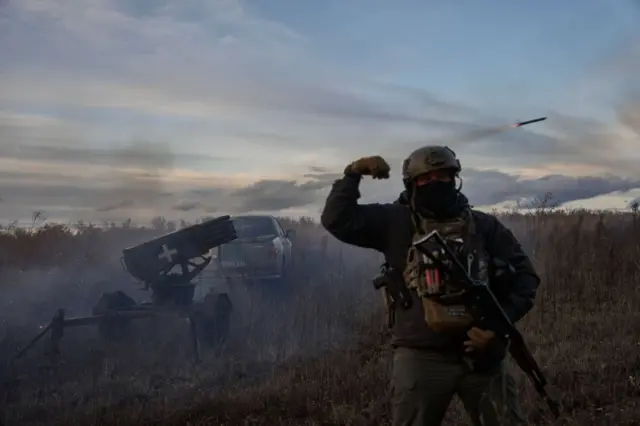 На передньому плані стоїть військовий у повному тактичному спорядженні з рацією, підсумками та зброєю, піднявши руку у переможному жесті. За кілька метрів позаду нього – встановлена імпровізована пускова установка, з якої щойно вилетів снаряд – його видно у вечірньому небі, легко затягнутому хмарами.