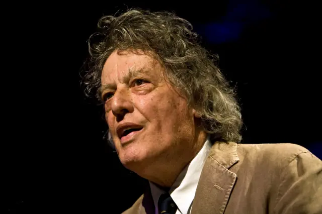 Sir Tom Stoppard has wavy, shoulder-length hair and is wearing a light brown corduroy jacket over a white shirt and a dark tie. The background is dark, suggesting a stage or event setting with focused lighting on Sir Tom.