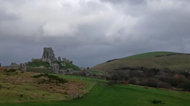 Los restos rocosos del castillo se encuentran en una colina en el horizonte, con nubes grises encima.