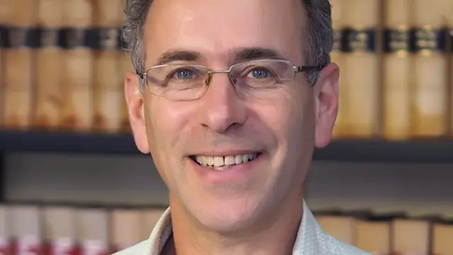 羅蘭·布朗直視鏡頭,他留著灰色短髮,戴著金屬框架眼鏡,背後的書架上放滿圖書