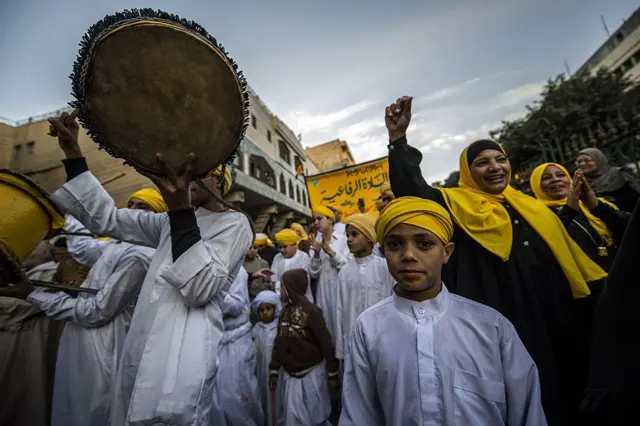 مجموعة من ارجال مصريين يرتدون طاقيات صفراء وجلاليب بيضاء وسيدات ترتدي غطاء رأس أصفر يسيرون في مجموعات ويحتفلون بالمولد النبوي الشريف، يحمل أحد الرجال دفاً.
