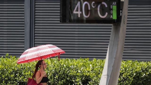 Mulher caminha sob guarda-chuva em dia de sol; grande painel mostra temperatura de 40ºC