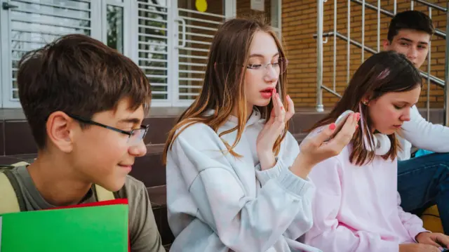 Una niña, rodeada de amigos, se maquilla frente a un espejo.