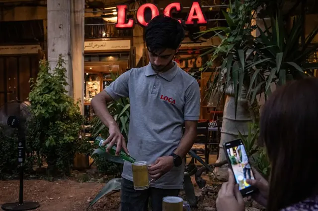 Üzerinde gri tişört olan bir genç bardağa bira döküyor. Bir kadın fotoğrafını çekiyor. 