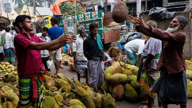 একটি বাজারে অনেক কাঁঠাল নিয়ে দাঁড়িয়ে আছেন কয়েকজন। লুঙ্গি ও শার্ট পরা একজন ব্যক্তি আরেকজনের দিকে একটি কাঁঠাল ছুড়ে দিচ্ছেন 