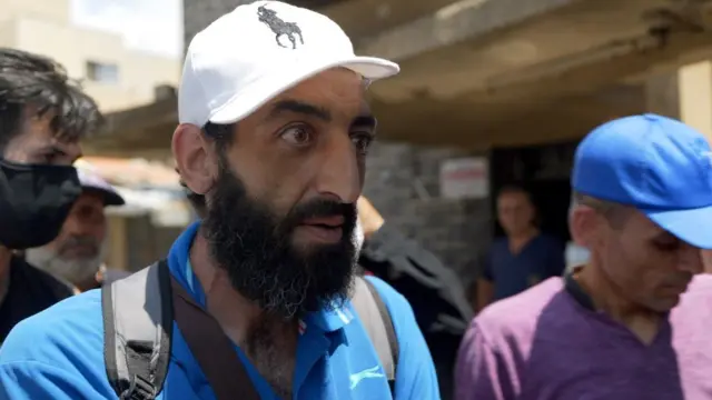 白いラルフローレンのキャップ、ストラップ付きのグレーのバックパック、青いポロシャツを着た男性が、カメラ外にいる記者と話している