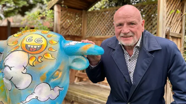 John Harris, con un abrigo azul y una camisa estampada, agachado junto a un toro de cerámica que compró en una subasta para el Birmingham Hospice.