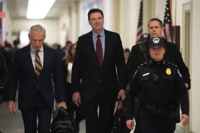  مدير مكتب التحقيقات الفيدرالي السابق، جيمس كومي، يقبل الإدلاء بشهادته أمام الكونغرس عام 2018، بخصوص قضية البريد الإلكتروني لهيلاري كلينتون وما إذا كان مستشارو حملة الرئيس ترامب تواطأوا مع الحكومة الروسية لتوجيه نتائج الانتخابات الرئاسية لعام 2016، ويظهر كومي مرتدياً سترة سوداء وقميصاً لونه أزرق فاتح وربطة عنق حمراء، وأمامه صابطة أمريكية مدججة بالسلاح وترتدي زي شرطة لونه أزرق.