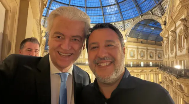 Sarışın bir erkek ile siyah saçlı bir erkek gülerek özçekim yapıyor. İhtişamlı bir yapı var arkalarında.