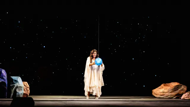 Atriz Regina Casé no centro de um palco. Ela veste uma longa túnica branca, segura com as mãos um globo terrestre pendurado por um fio que o deixa na altura de seu tórax. Ao fundo, projeções de estrelas em um fundo preto, imitando o espaço