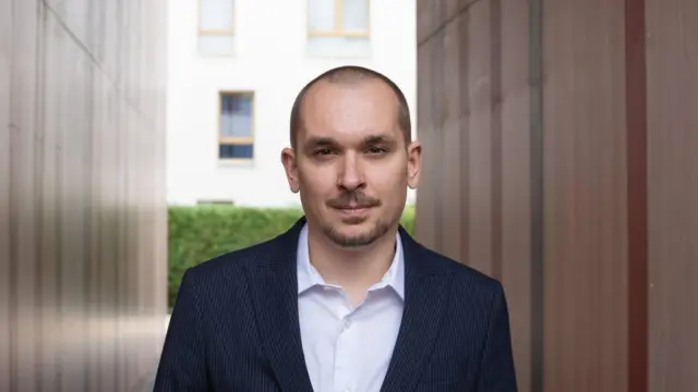 Журналист и бывший пресс-секретарь Сергея Тихановского Сергей Беспалов в темном костюме в мелкую полоску и с расстегнутой белой рубашке на фоне двух параллельных стен и жилого дома на заднем плане