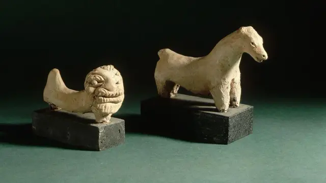 Duas estátuas levemente desgastadas, uma de um leão e outra de um cavalo, esculpidas em pedra bege clara e apoiadas sobre bases de madeira preta