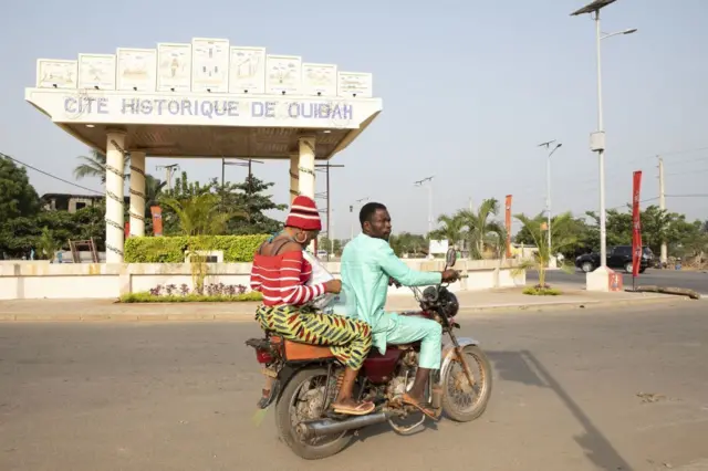 Un homme et une femme sur une moto circulant sur une route avec en arrière-plan un édifice sur lequel est écrit "Cité historique de Ouidah".