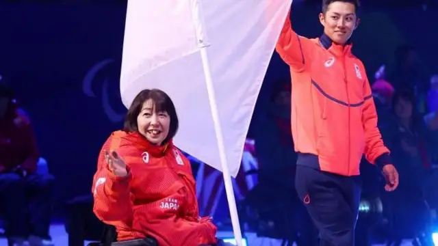 上半身赤系のウェアを着た2人が笑顔で行進している。2人の間には日本の国旗がある。小川は車いすに座り、右手を横に向けて伸ばし、その方向を見ている