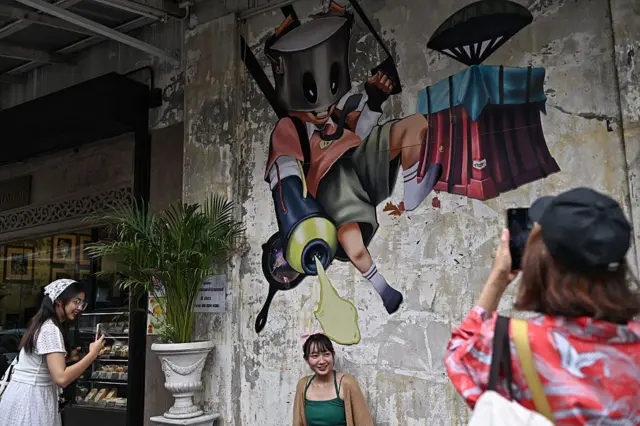 Visitors take pictures with art murals along Song Wat road in Bangkok on May 21, 2025. (Photo by Lillian SUWANRUMPHA / AFP) / RESTRICTED TO EDITORIAL USE - MANDATORY MENTION OF THE ARTIST UPON PUBLICATION - TO ILLUSTRATE THE EVENT AS SPECIFIED IN THE CAPTION (Photo by LILLIAN SUWANRUMPHA/AFP via Getty Images)