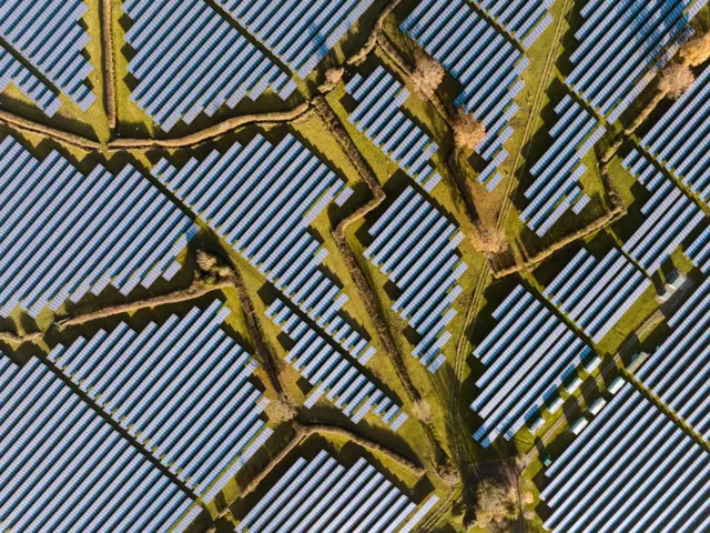 Uma vista aérea mostra fileiras de painéis solares dispostos em padrões geométricos, atravessados ​​por caminhos ou canais estreitos. As formas repetidas criam uma paisagem texturizada, semelhante a uma grade.