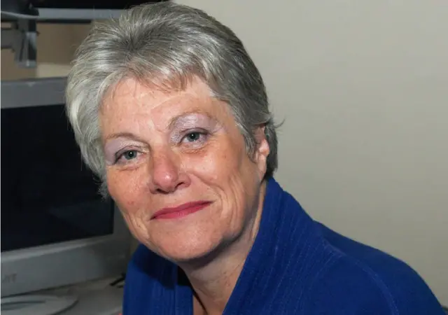 Christina Strang tem cabelos grisalhos curtos, está vestindo uma roupa azul e sentada em frente a um monitor de computador. O plano de fundo inclui parte do monitor e uma parede bege