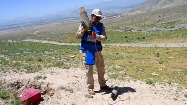 تصویر یک کارمند ماین‌پاک که یک ماده منفجر ناشده شبیه گلوله آوان را به شانه اش گرفته. این کارمند در یک دشت سبز دیده می شود.او واسکت آبی و کلاه خاکی به تن دارد. 