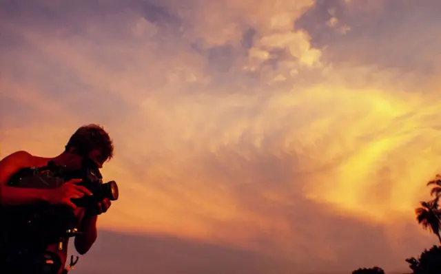A imagem mostra uma pessoa operando uma câmera de vídeo profissional ao ar livre durante o pôr do sol. Em silhueta contra um céu alaranjado e repleto de nuvens iluminadas, o cinegrafista aparece concentrado na filmagem, enquanto a luz quente do entardecer domina a composição. Ao fundo, a copa de uma palmeira reforça a atmosfera tropical da cena.