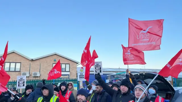 Se ve a varias personas, en su mayoría hombres, de pie junto a una valla de acero ondeando pancartas y banderas rojas con el nombre y el logotipo del sindicato Unite. Todos los hombres llevan chaquetas y muchos llevan capuchas o gorros de lana.