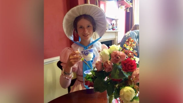 Una mujer con un gran sombrero estilo Regencia y un lazo azul bajo la barbilla. Está sentada a una mesa con un vestido rosa y sostiene una taza de té azul y blanca. Sobre la mesa, frente a ella, hay un jarrón con rosas.