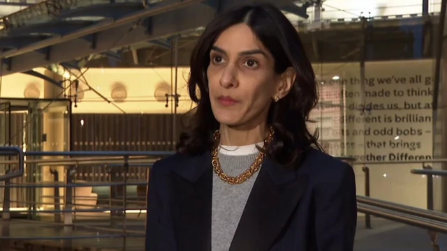 Priya Dogra, dyrektor generalna Channel 4, przed londyńską siedzibą stacji. Dogra to kobieta w średnim wieku, o czarnych włosach i brązowych oczach. Ma na sobie granatową marynarkę, jasnoszary sweter, złotą kolię i kolczyki. W tle ściana budynku wykonanego ze szkła i stali. 
