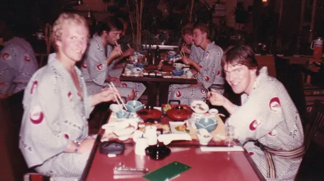 Taken in 1981, this photograph shows Bjørn-Eirik Olsen (left) seated at a table filled with an array of traditional Japanese dishes. Dressed in an elegant kimono and holding wooden chopsticks, Bjørn-Eirik faces the camera with a warm smile. Across the table sits a young man, Leif Inge Karlsen, later founder of Hydrotech Salmon Farm, wearing an identical kimono, sharing the moment and smiling towards the camera. Other young men wearing the same kimonos occupy similar tables in the background.
