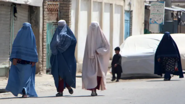 Burka giyen üç kadın önde yürüyor. Soldan sağa üzerlerinde açık mavi, lacivert ve bej rengi burka giyiyorlar. Giysiler kadınların gözleri dahil her yerini kapatıyor. Arkada siyah burkalı başka bir kadın yanında bir erkek çocuk ile yürüyor. Sokakta üzeri örtülü bir araç ve Peştuca tabelalar var.