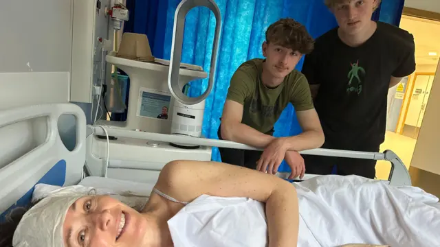 Dr Hard smiling while recovering in a hospital bed in London with her sons Callum and Oliver by her side.