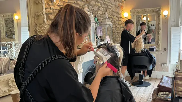 En una peluquería, dos jóvenes peluqueros vestidos de negro cortan el pelo. Sus clientas llevan capas negras. En las paredes cuelgan espejos con marcos ornamentados de color crema, lámparas redondas encendidas y una chimenea junto a una pared de piedra.