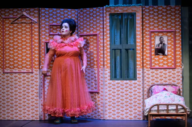 El personaje de Chula, vestida de naranja, frente a una escenografía sencilla que incluye el retrato de un hombre y una cama demasiado pequeña para el personaje