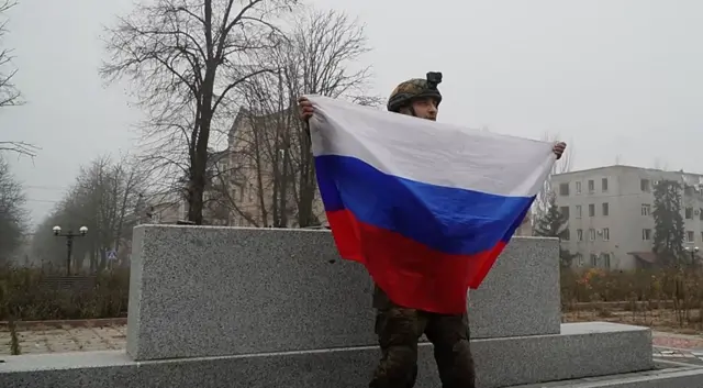 Российский военный с развернутым российским флагом в руках на фоне разбитого войной украинского городка