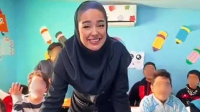 تصویر ویدیو معلم زن در حالی که به دوربین نگاه می‌کند و بچه‌های کلاسش پشت سرش دیده می‌شوند