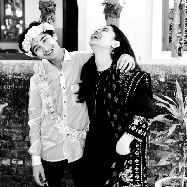 A black‑and‑white photo showing a teenage boy and a woman standing close together outdoors, one with an arm around the other. He is smiling, and she is laughing, her head back in the air. He is wearing a light-colored button‑up shirt and a long floral garland draped across the torso. She is wearing a dark, patterned outfit with embroidered details on the sleeves and a matching draped fabric. They stand in front of a textured brick wall, with plants and parts of a building visible in the background.