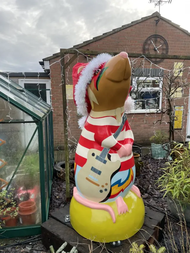 Imagen independiente de la escultura de la rata gigante rayada. Está colocada sobre un pedestal amarillo y luce un elaborado maquillaje de ojos.