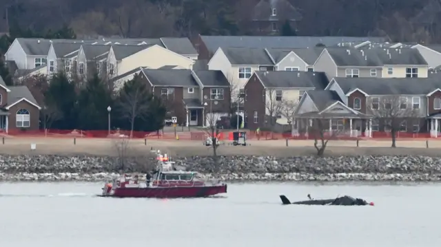Упавший вертолет торчит из воды, рядом с ним катер спасателей, на заднем плане на берегу двухэтажные дома 
