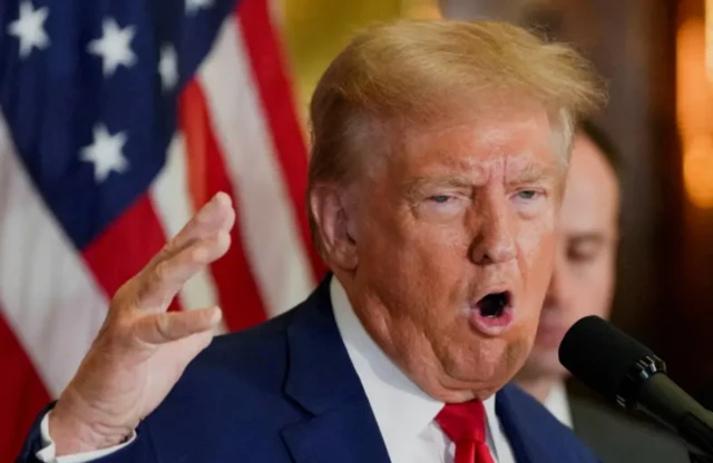 Donald Trump com a boca aberta em frente a um microfone e com a bandeira dos Estados Unidos ao fundo