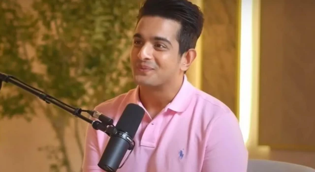 Ranveer Allahbadia during his podcast. He is wearing a light pink polo shirt and is sitting in front of a mic in a warmly lit studio with yellow interiors. 