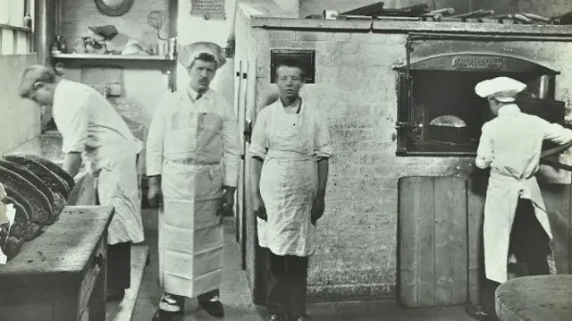 Scena w piekarni; jeden chłopiec wyjmuje bochenek chleba z pieca, drugi pozuje z instruktorem, a trzeci zajmuje się ciastem.