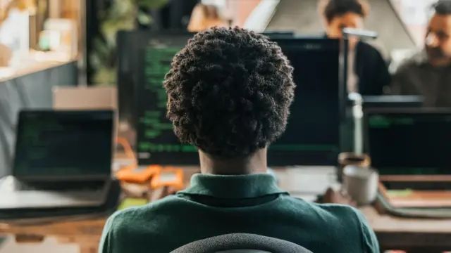 Mężczyzna z kręconymi włosami koduje na komputerze stacjonarnym, siedząc na krześle w biurze.