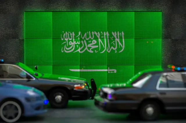 Carros passando em frente a tela com bandeira da Arábia Saudita