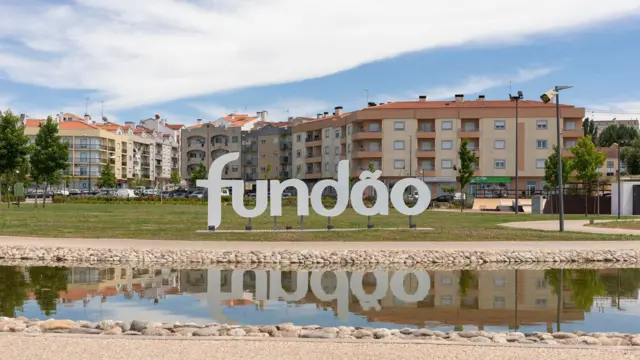 Um letreiro com o nome da cidade Fundão, em frente a edifícios baixo e com um curso de água em primeiro plano