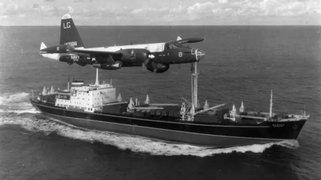 একটি মার্কিন টহল বিমান সমুদ্রে একটি পণ্যবাহী জাহাজের পাশ দিয়ে উড়ছে। জাহাজটির একটি উঁচু মাস্তুল এবং সামনের অংশে একটি কালো রঙের রেখা দেখা যাচ্ছে 