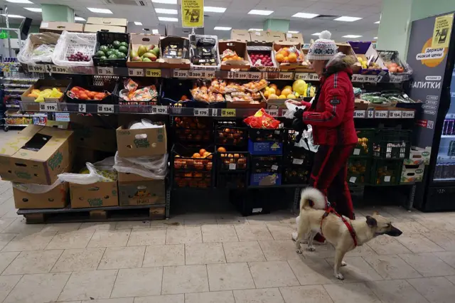 Uma mulher com um casaco de inverno vermelho e um cachorrinho na coleira caminham em direção à seção de frutas do supermercado.