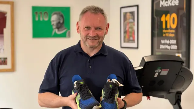 Gary McKee lleva un polo azul y unas zapatillas deportivas. Sonríe y tiene el pelo corto y la barba grises.