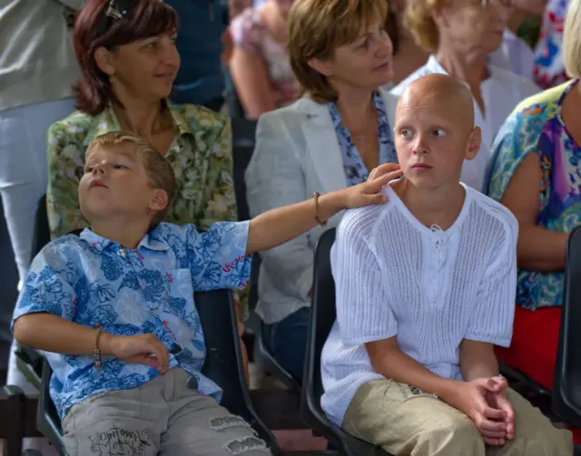Un niño rubio tiene su mano sobre el hombro de un niño que ha perdido su cabello, durante lo que pareciera ser un evento