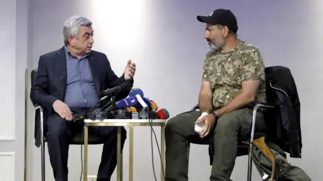Sarkisyan ve Paşinyan karşılıklı oturuyor. Sarkisyan'ın üzerinde takım elbise ve önünde mikrofonlar var. Paşinyan kamuflaj desenli tişört giyiyor, siyah bir şapka takıyor. Paşinyan'ın sağ eli sargılı.